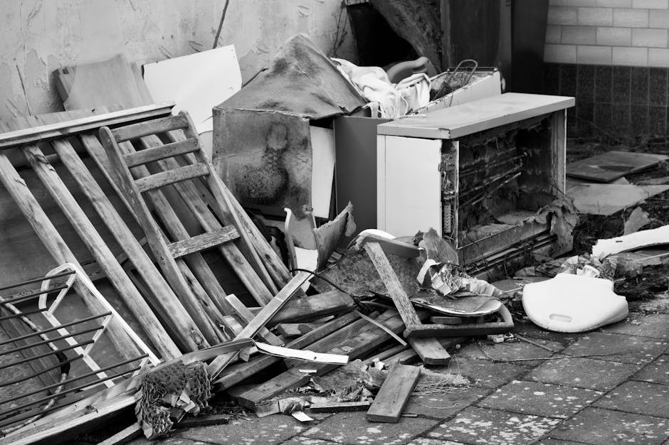 A pile of discarded indoor items and debris situated outdoors on a paved surface near a brick and tile wall. Prominent in the scene is a wooden garden trellis or rack, leaning against the left side, w