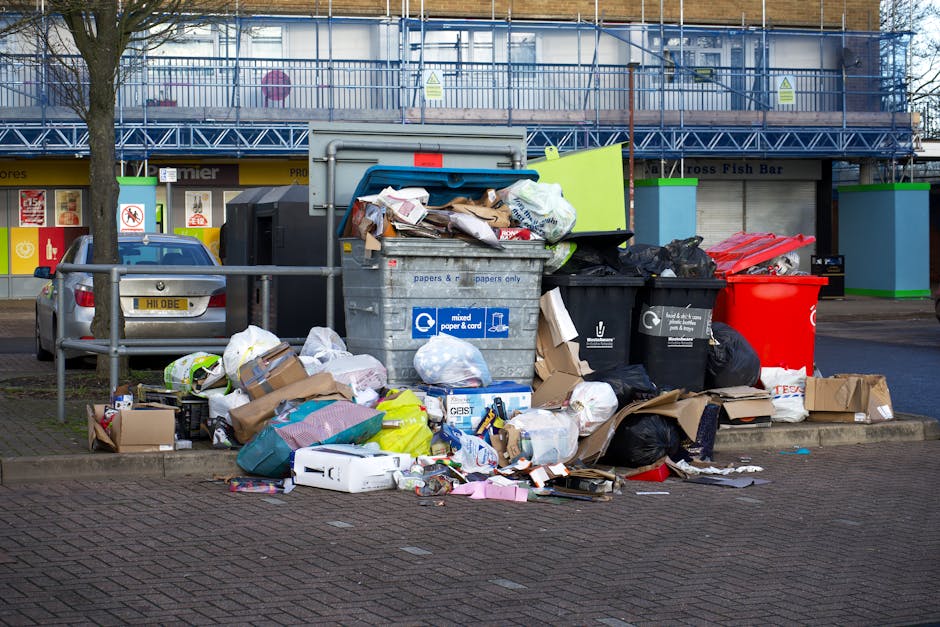 A pile of discarded indoor items and debris situated outdoors on a paved surface near a brick and tile wall. Prominent in the scene is a wooden garden trellis or rack, leaning against the left side, w