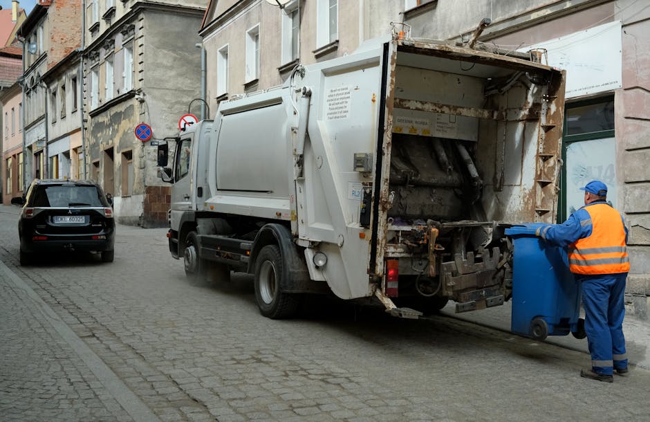 A worker in a yellow and red high-visibility vest is operating a large red waste collection vehicle on a paved street. The back of the vehicle is open, revealing mechanical components and a loading ar