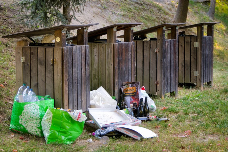 A person dressed in dark clothing, including yellow rubber boots, is walking along a dirt path outdoors during daylight, carrying two large, white plastic bags filled with rubbish or waste materials.