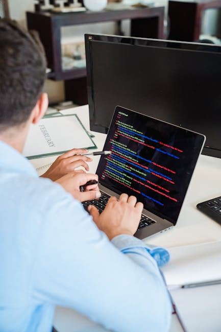 A person wearing a light blue shirt is sitting at a desk, working on a laptop with a screen displaying lines of colorful computer code. The individual's right hand is resting on the keyboard, while th